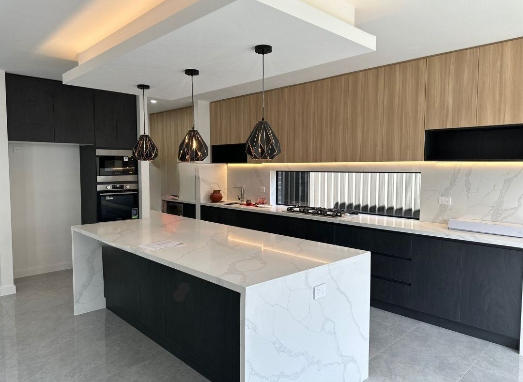 Long galley kitchen with a mitred waterfall stone island, oak and black overhead cupboards, three black pendant lights and a window splashback.